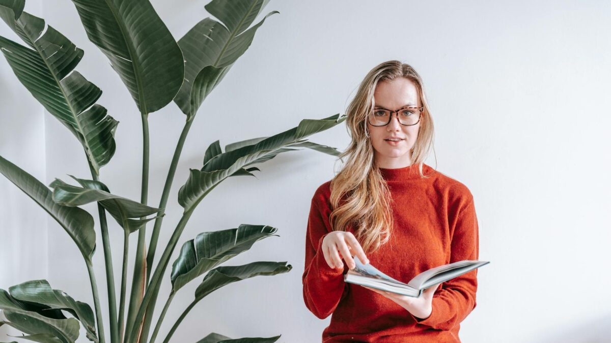 A young woman in a sweater reads a book indoors beside a lush green plant.